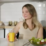 усміхнена жінка на кухні сидить за столом із тарілкою салату та фрешем